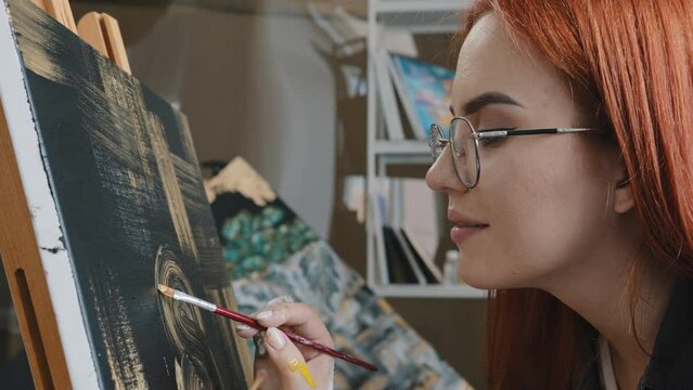 Side view close up red-haired 20s woman talented artist girl in glasses female painter paints picture on black background with golden oil acrylic lady painting on canvas learning studying art skill