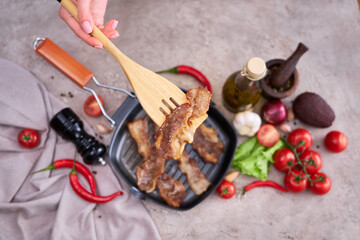 Fototapeta na wymiar fried bacon pieces in a skillet and vegetables at domestic kitchen stone or concrete table