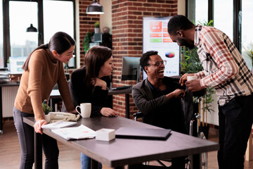 African american man with physical impairment having fun at work, taking break with workmates. Male wheelchair user suffering from chronic disability laughing with business partners.