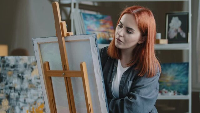 Portrait pensive red-haired Caucasian woman paints in room thinking about idea create picture ponder think drawing hobby talented girl painting on canvas with acrylic contemporary art work in studio