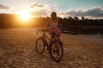 Fototapeta na wymiar Attractive red-haired young woman rides a bicycle on the beach in shorts. Evening. Sunset. sand
