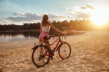 Fototapeta na wymiar Attractive red-haired young woman rides a bicycle on the beach in shorts. Evening. Sunset. sand