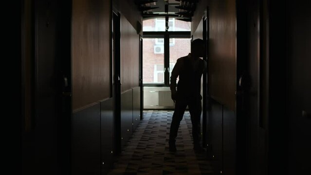 silhouette of unrecognizable man leaves hotel room in hallway. businessman in trousers and vest walks forward along dark corridor, behind window creates light. concept of business trip