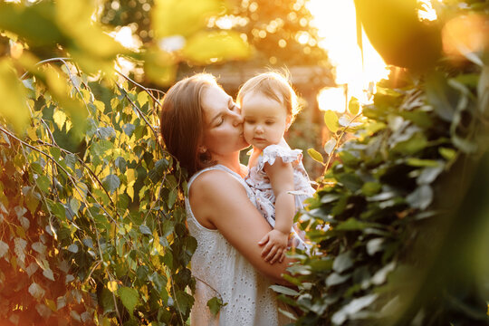 Happy young mother playing and kissing her little baby daughter on sunshine warm spring or summer day. Beautiful sunset light in the garden or in the park. Happy family concept.