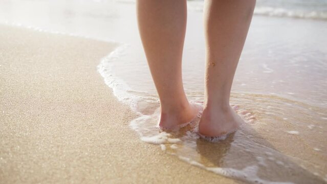 Woman feet walking barefoot on sandy beach of sea. Scenic summer background in slow motion. Beautiful golden sunlight reflecting from the wet beach surface.Woman standing in ocean waves on sandy beach