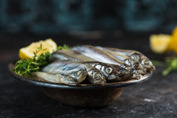 Pickled capelin with a slice of lemon and thyme on a metal plate on a dark background.