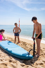 Fototapeta premium Three kids inflating a paddle surf on the beach