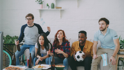 Naklejka premium Multi-ethnic group of friends sports fans with Argentinian flags watching football championship on TV together at home indoors and cheering up favourite team