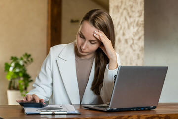 Fototapeta premium A lonely woman in a white jacket with a lot of debts, experiencing stress, counting finances, sitting at a table with papers, a laptop, doing calculations on a calculator, trying to make ends meet