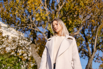 Stylish confident woman walking in the park in fall. Modern autumn outfit. Girl in a coat. Falling leaves, yellow background. Sunny warm day in the city. Good beautiful weather