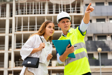 Male engineer in uniform and white hardhat pointing with finger while talking with woman in...