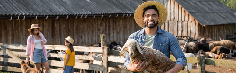 farmer in straw hat holding lamb and smiling at camera near family at corral, banner.