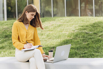 Portrait of focused student girl doing homework next to laptop computer. Teenage girl writing in notebook in front of laptop outdoors. Pretty girl studying at home education