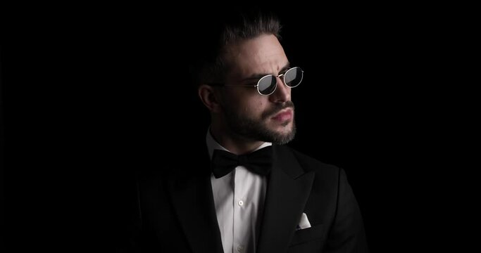 sexy unshaved businessman in tuxedo with bowtie and sunglasses looking to side, arranging tux, being confident and mysteriously disappearing in the dark