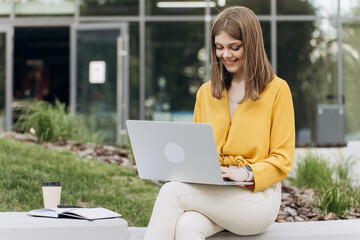 Attractive young female using laptop while sitting on bench outdoors in city. Pretty girl student preparing for classes in nature. Lady corporate start up office employee working outdoor.