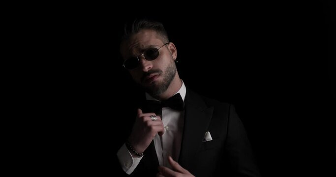 mysterious sexy groom rubbing palms, looking to side, adjusting tuxedo and sunglasses and crossing arms in the dark