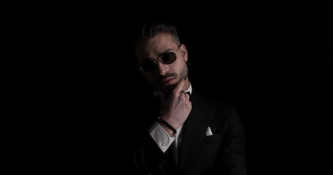 seductive young man in tuxedo touching and rubbing beard, fixing bowtie and sunglasses, looking to side, crossing arms in front of black background