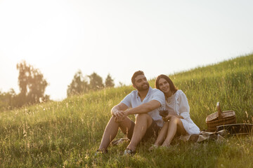 Fototapeta premium joyful couple sitting on green slope near wicker basket and looking away.