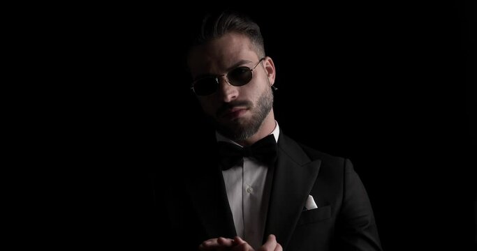 project video of elegant young businessman in tuxedo rubbing palms, looking to side and posing in the dark