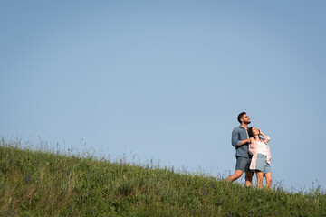 happy couple in denim shorts standing on green hill under blue sky and looking away.