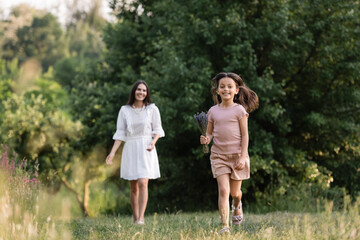 full length of cheerful girl with lavender bouquet running near mom on blurred background.