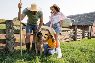 Naklejka na ściany i meble girl stroking cattle dog near parents with rakes and bowl on farm.