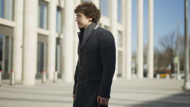 A young man with dark curly hair walks, looks up, stops, takes a picture on his phone. Against the backdrop of an office building. 