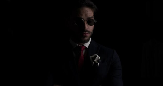 mysterious businessman pulling down sunglasses and looking over, looking to side and arranging tie while posing on black background