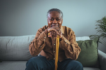Portrait of happy senior man smiling at home while holding walking cane. Old man relaxing on sofa...
