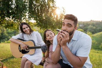 man playing harmonica near daughter with bouquet and wife with acoustic guitar. - obrazy, fototapety, plakaty