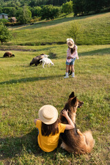 girl in straw hat sitting with dog while parents hugging near herd grazing in pasture.
