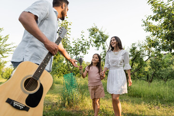 man with guitar outstretching hand near wife and daughter smiling outdoors.