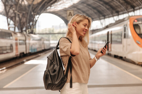 Pretty cute woman using her phone at the railway station . European fair-haired lady with black backpack keep hand on head 