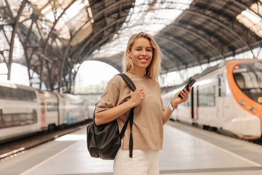 Attractive european woman look with smile at camera and holding phone . Blonde lady at railway station with black backpack 