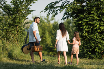 man with guitar and wicker basket smiling near family walking in countryside.