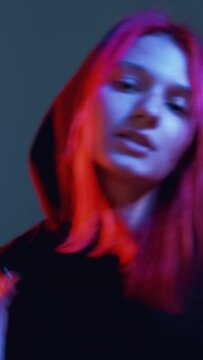 Vertical video. Neon light dance. Hip-hop night. Cyberpunk party. Red blue color funky redhead girl with in hoodie breakdancing on dark background.