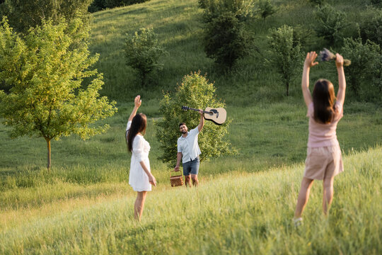 woman and girl waving hands to happy man with acoustic guitar on grassy slope.