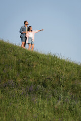 brunette woman pointing with hand while standing with man on green hill.