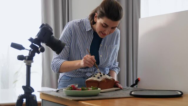 blogging, cooking and people concept - happy smiling female photographer or food blogger with camera, cake and brush working in kitchen at home