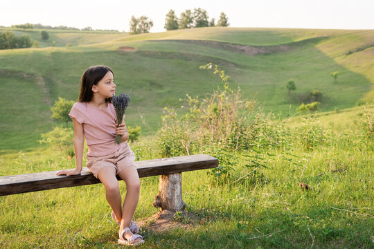 full length of girl with lavender flowers sitting on bench in countryside and looking away.