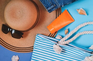 Summer travel 2022 lifestyle photography. Straw hat, sunglasses, striped bag, orange sunscreen tube and seashells on a blue background. Sea vacation, holidays concept, object photo