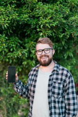 Happy bearded man showing blank smartphone screen over green leaves background - empty space for advertising and copy space