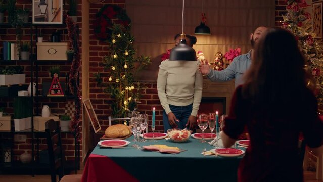 Interracial couple welcoming senior people and young woman at Christmas family dinner table. Festive people gathering at home to celebrate winter holiday while giving each other gifts.