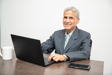 Portrait of happy senior Indian businessman working on a laptop in office. Elderly mature gray haired asian corporate male using computer.