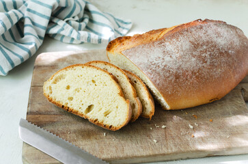 Slices of fresh baked bread on a wooden cutting board with a knife and a dish towel on a light background. Rustic style. The concept of homemade bread. Horizontal orientation.