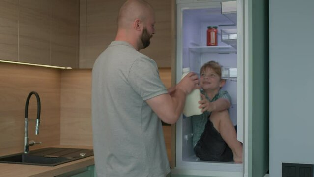 Father playing with son sitting in the fridge and giving him milk bottle. Family having fun at home.