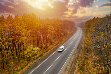 white car driving on the asphalt road through the autumn forest at sunset. travel and transportation concept