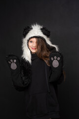 Portrait of a cheerful cute teenage girl with long black hair, dressed in a panda fur hat, posing on a black studio background. A comical image. Advertisement. original children's clothing.