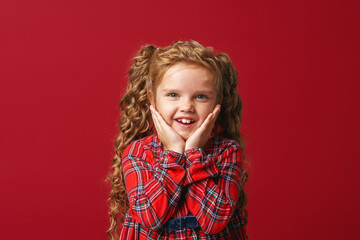 Portrait of a charming little girl, enthusiastically rejoicing, standing on a red background in the studio. The child is emotionally excited and looks at the camera. Advertising