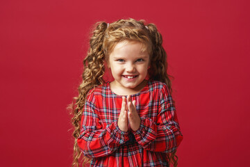 cunning little girl, 7 years old, plotting something bad, meanness, a child with an insidious expression looks at the camera standing on a red background in the studio.